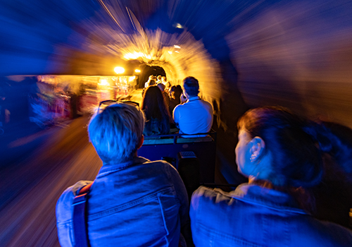 Menschen sitzen in einem pffenen Waggon und fahren durch einen beleuchteten Tunnel